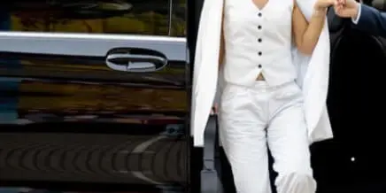 A professional chauffeur assisting a woman in a white suit step out of a luxury black car.