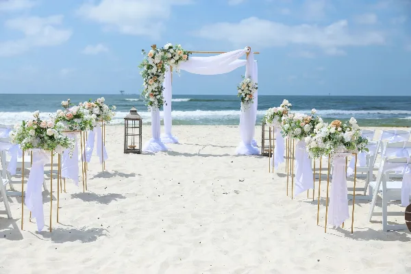 A beach wedding ceremony setup with a floral arch, white drapery, and floral aisle stands on the sand.