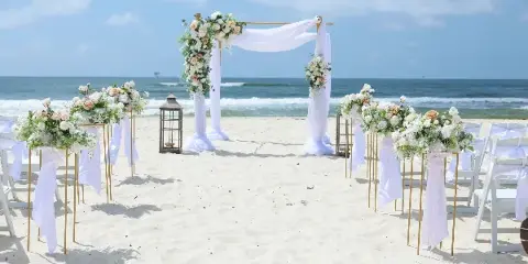A beach wedding ceremony setup with a floral arch, white drapery, and floral aisle stands on the sand.