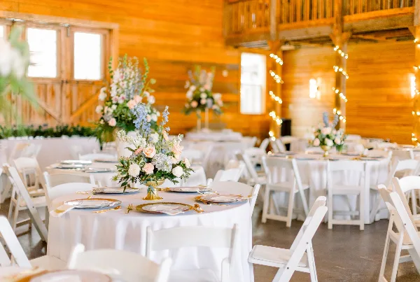 Interior of a rustic wooden barn venue with round tables, white linens, floral centerpieces, and string lights set up for a wedding reception.