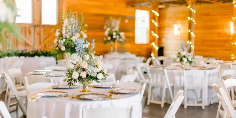 Interior of a rustic wooden barn venue with round tables, white linens, floral centerpieces, and string lights set up for a wedding reception.