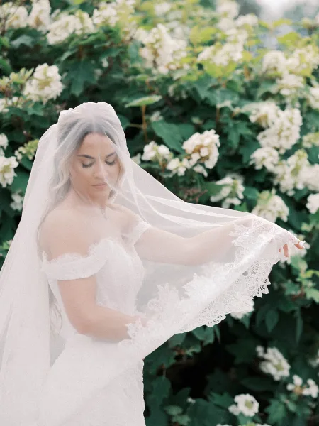 A bride in a white lace dress and veil standing in front of a flowering bush.
