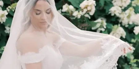 A bride in a white lace dress and veil standing in front of a flowering bush.