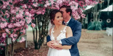 Bride and groom embracing outdoors beneath a flowering pink tree.