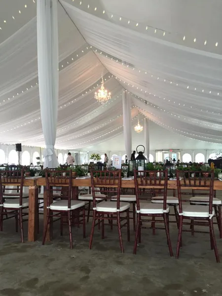 Interior of a white wedding tent featuring draped ceiling fabric, string lights, and wooden dining chairs.