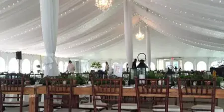 Interior of a white wedding tent featuring draped ceiling fabric, string lights, and wooden dining chairs.