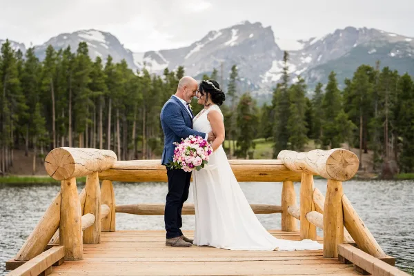 A bride and groom standing on a wooden dock with a mountain and forest background.