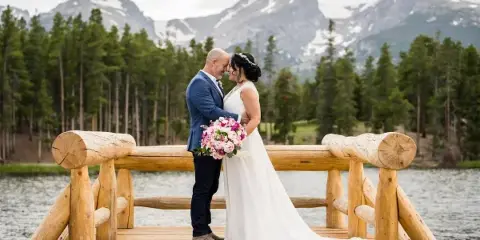 A bride and groom standing on a wooden dock with a mountain and forest background.