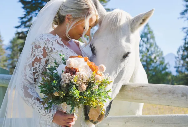 Bride in lace gown and veil holding a bouquet while touching a white horse near a wooden fence with mountains in the background.