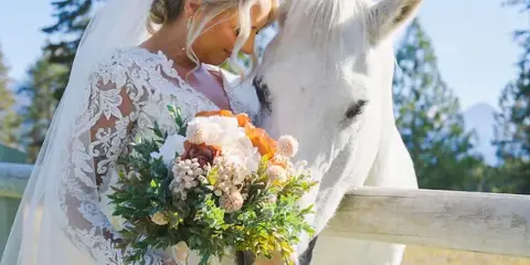 Bride in lace gown and veil holding a bouquet while touching a white horse near a wooden fence with mountains in the background.