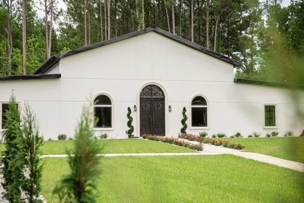 Exterior view of a white stucco wedding venue building with an arched entryway, manicured lawn, and surrounding pine trees.