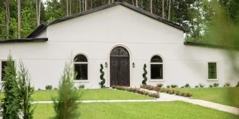 Exterior view of a white stucco wedding venue building with an arched entryway, manicured lawn, and surrounding pine trees.