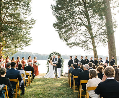 A bride and groom exchanging vows at an outdoor wedding ceremony on a lawn overlooking a river, with guests seated in rows of chairs.