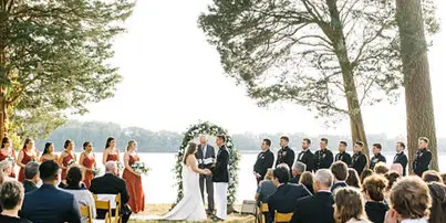 A bride and groom exchanging vows at an outdoor wedding ceremony on a lawn overlooking a river, with guests seated in rows of chairs.