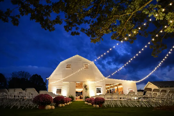 A white barn wedding venue at dusk with rows of chairs, floral centerpieces, and string lights.
