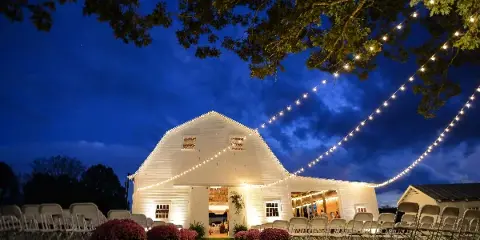 A white barn wedding venue at dusk with rows of chairs, floral centerpieces, and string lights.
