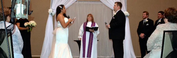 A wedding officiant in white and purple vestments stands at an altar with a bride and groom during a ceremony.