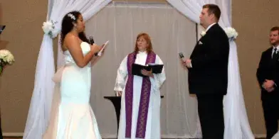 A wedding officiant in white and purple vestments stands at an altar with a bride and groom during a ceremony.