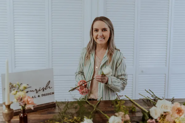 A woman florist arranging fresh flowers and greenery at a work table with a Flourish Floral & Design sign.