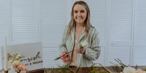 A woman florist arranging fresh flowers and greenery at a work table with a Flourish Floral & Design sign.