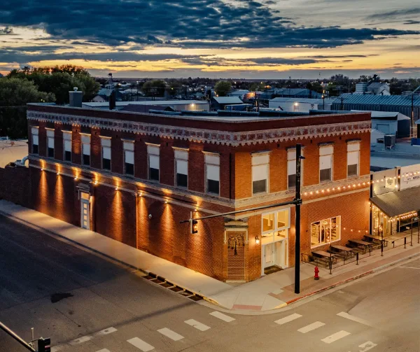 Aerial view of a two-story red brick building at a street corner in downtown Yuma, Colorado, illuminated at dusk with string lights and outdoor seating.
