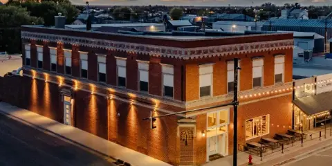 Aerial view of a two-story red brick building at a street corner in downtown Yuma, Colorado, illuminated at dusk with string lights and outdoor seating.