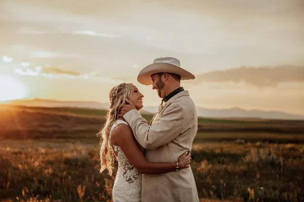 A bride and groom embracing in a grassy field at sunset with mountains in the distance.