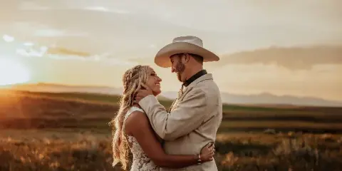 A bride and groom embracing in a grassy field at sunset with mountains in the distance.