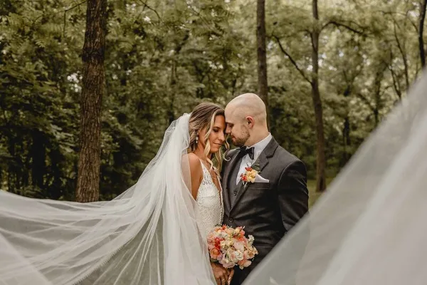 A bride and groom standing together in a wooded area, with the bride's veil blowing in the wind.