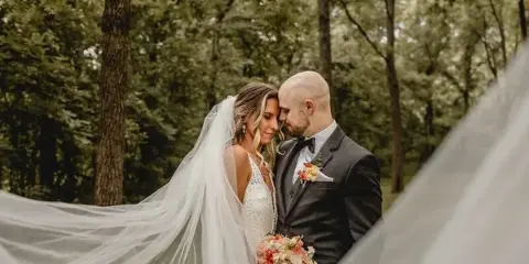 A bride and groom standing together in a wooded area, with the bride's veil blowing in the wind.