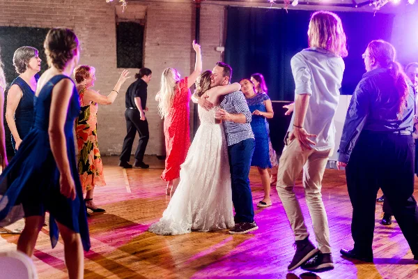 A bride and groom dancing at a wedding reception with guests under purple and pink dance floor lighting.