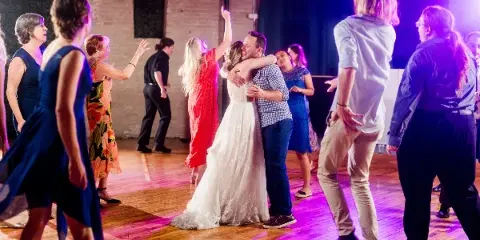 A bride and groom dancing at a wedding reception with guests under purple and pink dance floor lighting.