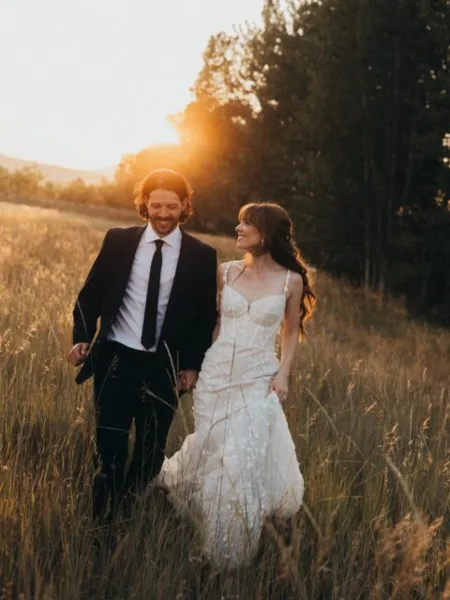 A bride and groom walking through a tall grass field during golden hour.