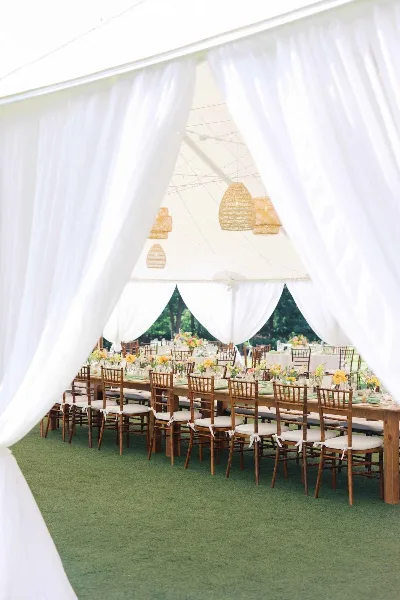 A luxury wedding reception setup inside a white tent with drapery, featuring long wooden tables, Chiavari chairs, and floral centerpieces.