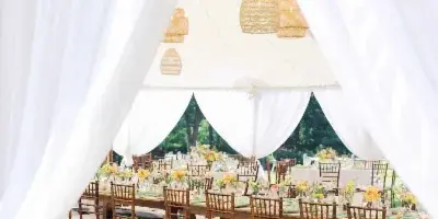 A luxury wedding reception setup inside a white tent with drapery, featuring long wooden tables, Chiavari chairs, and floral centerpieces.