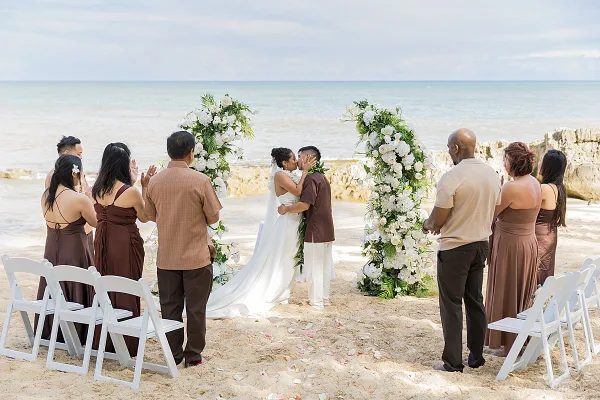 A bride and groom exchanging vows under a floral arch on a sandy beach with guests seated in white chairs.