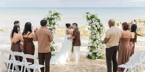 A bride and groom exchanging vows under a floral arch on a sandy beach with guests seated in white chairs.