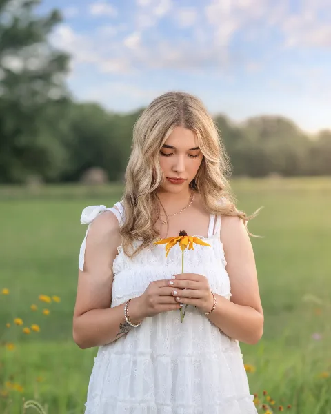 A young woman with blonde hair wearing a white dress stands in a grassy field holding a yellow flower.
