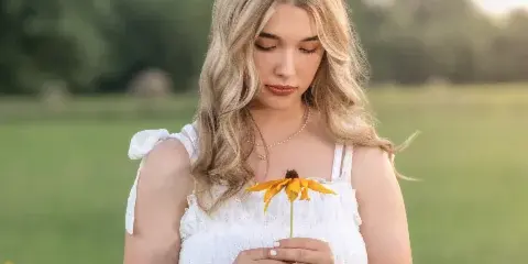 A young woman with blonde hair wearing a white dress stands in a grassy field holding a yellow flower.