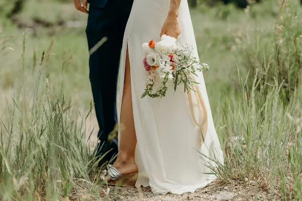 A bride in a white gown holding a bouquet standing next to a groom in a dark suit in a grassy field.