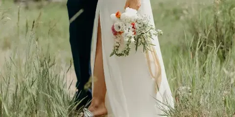 A bride in a white gown holding a bouquet standing next to a groom in a dark suit in a grassy field.