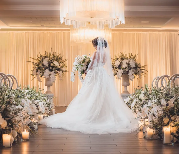 A bride in a white gown stands in a decorated wedding venue featuring large floral arrangements, candles, and elegant drapery.