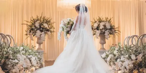 A bride in a white gown stands in a decorated wedding venue featuring large floral arrangements, candles, and elegant drapery.