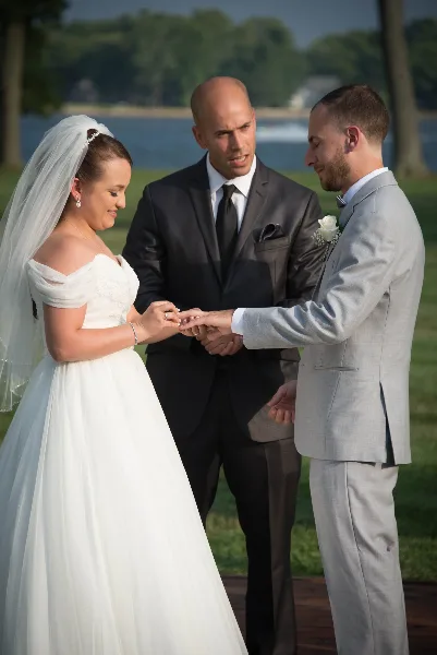 A wedding officiant in a dark suit stands between a bride and groom during an outdoor ceremony, overseeing the exchange of rings.