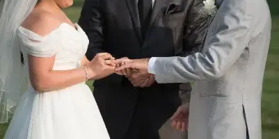 A wedding officiant in a dark suit stands between a bride and groom during an outdoor ceremony, overseeing the exchange of rings.