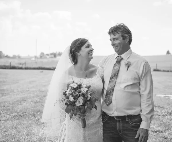 A bride and a man in a dress shirt and tie standing together outdoors in a field, smiling at each other.