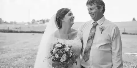 A bride and a man in a dress shirt and tie standing together outdoors in a field, smiling at each other.