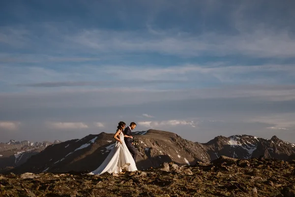 A bride and groom standing on a rocky mountain summit with a vast mountain range in the background.