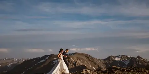 A bride and groom standing on a rocky mountain summit with a vast mountain range in the background.