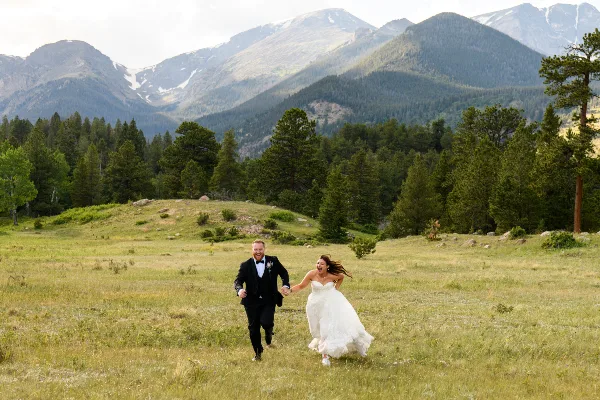 A bride and groom running hand-in-hand through a grassy meadow with a backdrop of pine trees and snow-capped mountains.